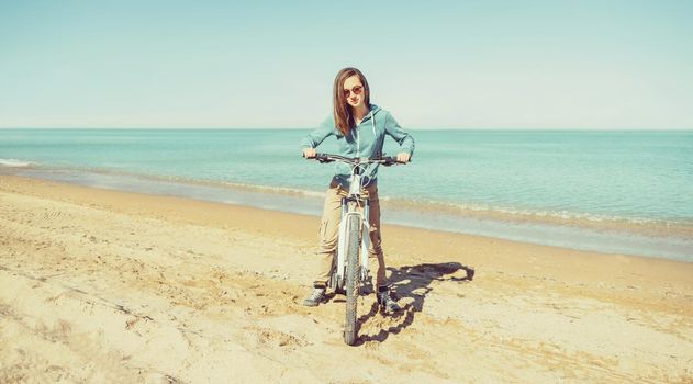 Beautiful girl in sunglasses standing with bicycle on sand beach on background of sea in summer