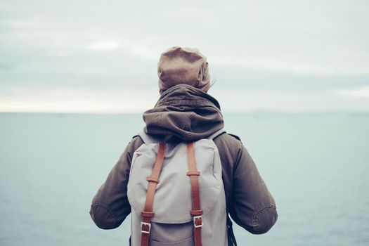 Unrecognizable traveler wearing in warm clothing with backpack enjoying view of sea, rear view