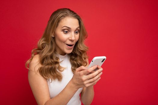Image of a beautiful shocked young blonde woman posing isolated over red wall background using mobile phone