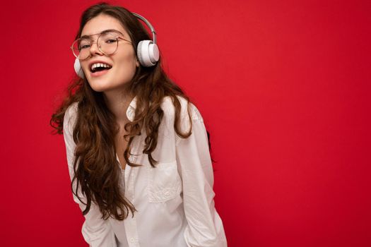 Photo of attractive positive smiling young brunet woman wearing white shirt and optical glasses isolated over red background wearing white wireless bluetooth headsets listening to music and having fun. empty space
