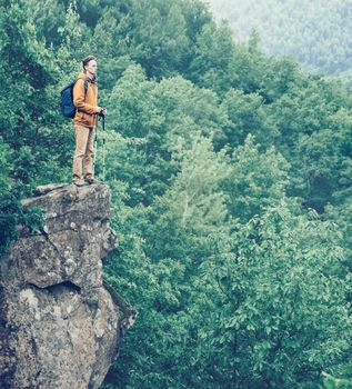 Hiker with backpack and trekking poles standing on edge of cliff and enjoying view of nature in summer outdoor