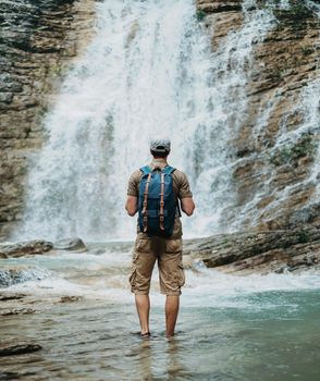 Unrecognizable traveler young man with backpack standing in river and looking at waterfall in summer outdoor, rear view