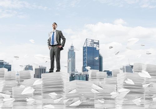 Confident businessman in suit standing on pile of documents among flying paper planes with cityscape on background. Mixed media.