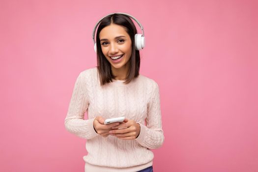 Photo shot of beautiful joyful smiling young female person wearing stylish casual outfit isolated over colorful background wall wearing white bluetooth wireless earphones and listening to music and using mobile phone looking at camera.