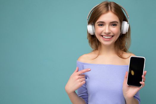 Attractive happy young blond woman wearing blue crop top isolated over blue background wall holding and showing mobile phone with empty display for mockup and wearing bluetooth headphones looking at camera. copy space