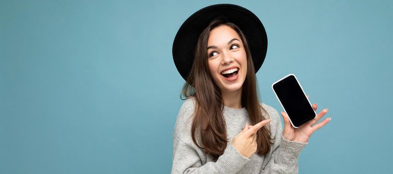 Panoramic Closeup of charming young happy woman wearing black hat and grey sweater holding phone looking to the side pointing finger at screen isolated on background.Mock up, cutout