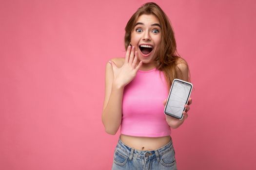 Photo of beautiful surprised amazed smiling young woman good looking wearing casual stylish outfit standing isolated on background with copy space holding smartphone showing phone in hand with empty screen display for mockup pointing at gadjet looking at camera.