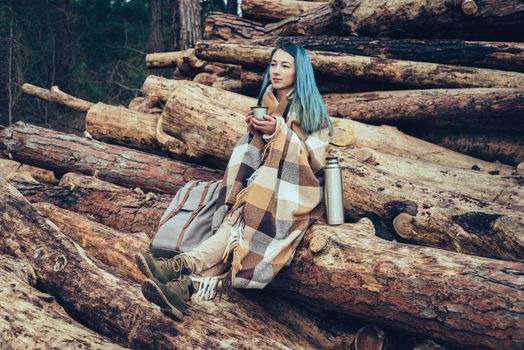 Young woman wrapped in plaid resting with cup of tea on tree trunks outdoor
