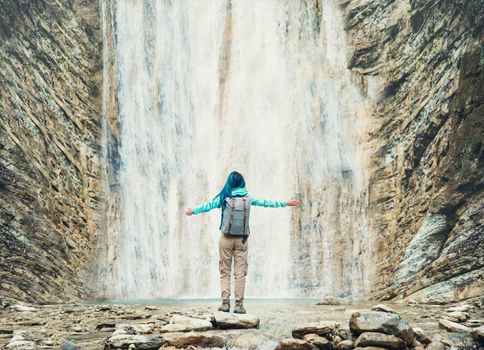 Happy explorer young woman with backpack standing with raised arms in front of beautiful waterfall