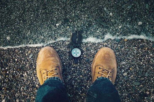 Compass on pebble shore near legs of traveler man outdoor. Point of view shot.