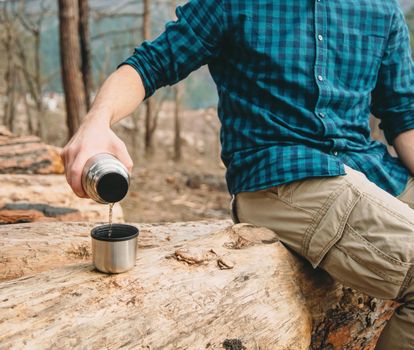 Unrecognizable hiker young man pouring tea from thermos to cup in the forest outdoor