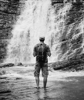 Unrecognizable traveler young man with backpack standing in river and looking at waterfall in summer outdoor, rear view. Monochrome image