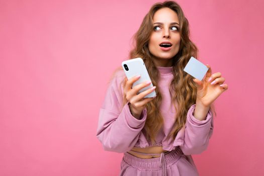 Photo of attractive thoughtful with open mouth young blonde curly woman wearing pink clothes isolated over pink background using mobile phone making payment using credit card looking at to the side and afraiding of smth.