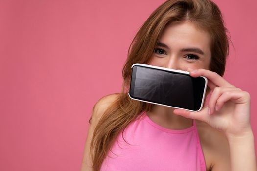 Photo of beautiful positive young blonde woman wearing pink top poising isolated on pink background with empty space holding in hand and showing mobile phone with empty display for cutout looking at camera.