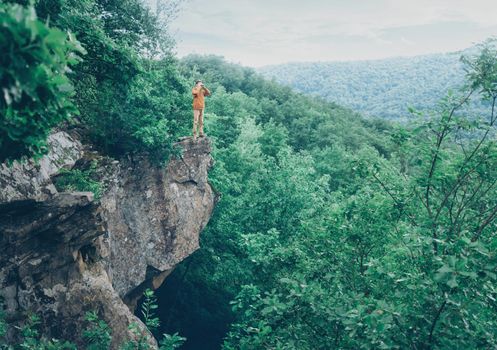 Traveler young man standing on edge of cliff and looking in binoculars into the distance in summer outdoor