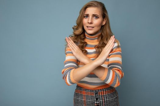 Photo of young attractive upset angry dissatisfied blonde woman with sincere emotions wearing striped sweater isolated over blue background with copy space and showing no gesture and stop sign with crossed hands. Negative concept.
