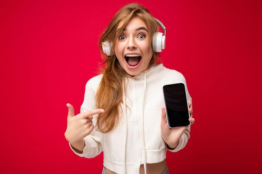 Beautiful positive amazed young woman wearing stylish casual outfit isolated on background wall holding and showing mobile phone with empty display for mockup wearing white bluetooth headphones listening to music and having fun looking at camera.