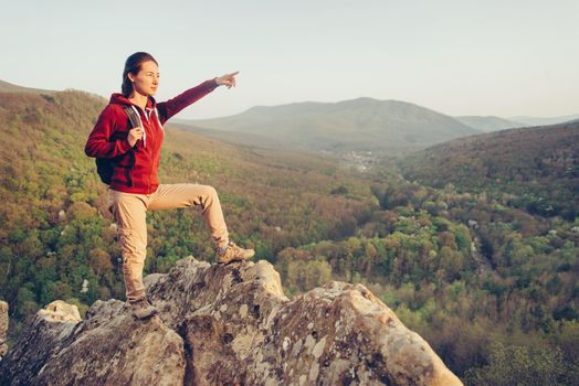 Hiker young woman with backpack standing on peak of rock on background of mountains and pointing into the distance in summer outdoor