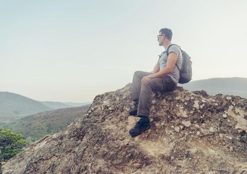 Hiker young man with backpack sitting on peak of rock on background of mountains and enjoying view of landscape in summer outdoor