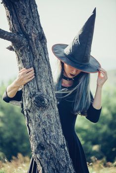 Beautiful young woman in costume of witch standing near the tree outdoor. Theme of Halloween and magic