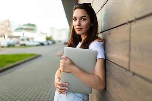 Close-up portrait of nice-looking charming attractive lovely fascinating cheerful cheery straight-haired brunet lady looking at camera in the street with laptop computer and glasses in hands.