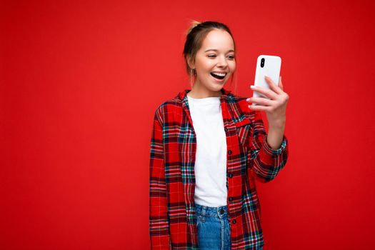 Photo shot of attractive positive good looking young woman wearing casual stylish outfit poising isolated on background with empty space holding in hand and using mobile phone messaging sms looking at smartphone display screen.