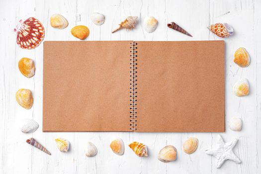 Open notepad with sea shells on white wooden background, top view