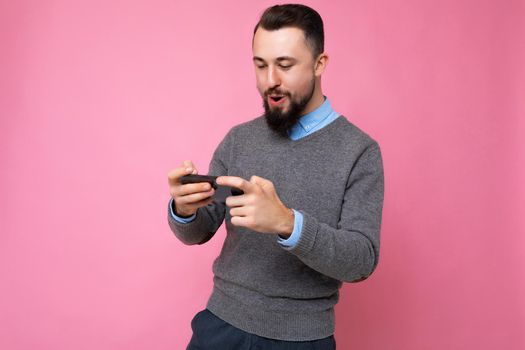Shot of happy handsome young brunette unshaven man with beard wearing everyday grey sweater and blue shirt isolated on background wall holding smartphone playing games via mobile phone looking at phone screen display and having fun.