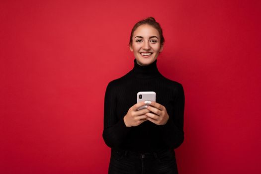 Photo shot of attractive positive good looking young woman wearing casual stylish outfit poising isolated on background with empty space holding in hand and using mobile phone messaging sms looking at camera.