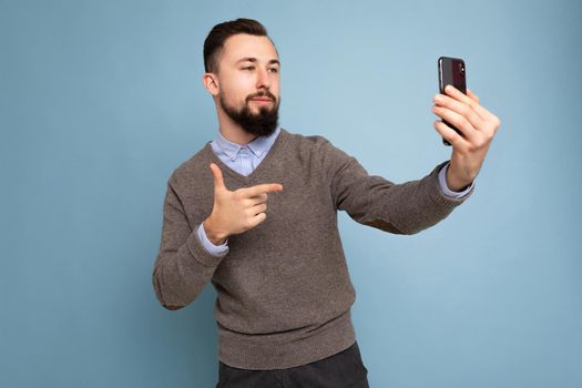Photo of positive handsome young brunette unshaven man with beard wearing casual grey sweater and blue shirt isolated on pink background wall holding smartphone taking selfie photo looking at mobile phone screen display.
