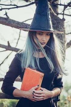 Dark young witch standing with book in the forest, looking at camera. Space for text on book