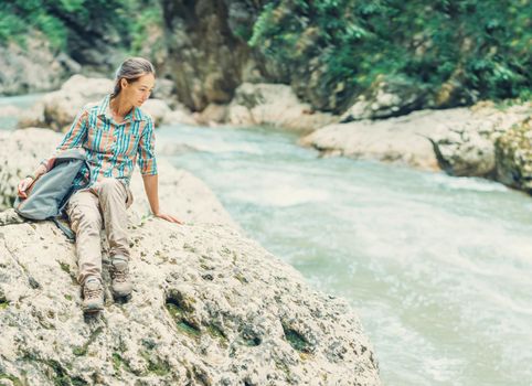 Traveler young woman with backpack sitting on stone and looking on mountain river in summer