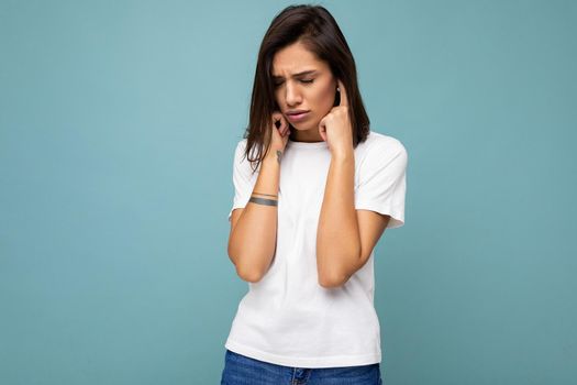 Portrait of sad upset young beautiful brunette woman with sincere emotions wearing casual white t-shirt for mockup isolated on blue background with empty space and covering ears with fingers.