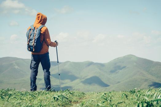 Unrecognizable traveler young man with backpack and trekking poles looking at mountains in summer outdoor. Free space in right part of the image