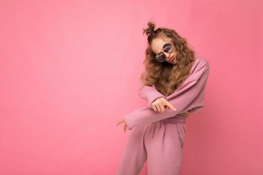 Shot of beautiful happy young dark blonde curly woman isolated over pink background wall wearing casual pink sport clothes and stylish sunglasses looking at camera and dancing. copy space