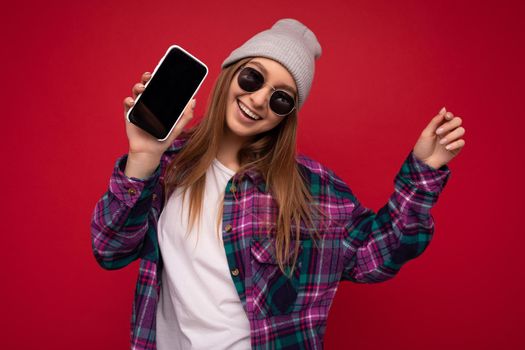 Photo of beautiful positive young blonde woman wearing stylish purple shirt and casual white t-shirt grey hat and sunglasses isolated over red background holding in hand and showing mobile phone with empty screen for mockup looking at camera dancing and having fun.
