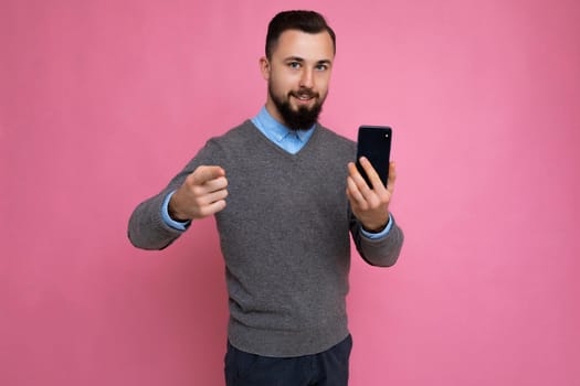 Good looking happy cool young brunette unshaven man with beardwearing stylish grey sweater and blue shirt standing isolated over pink background wall holding smartphone and using phone looking at camera pointing finger at camera.