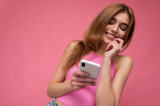 Photo of good looking young blonde woman wearing pink top poising isolated on pink background with empty space holding in hand and using mobile phone messaging sms looking at gadjet display and thinking.