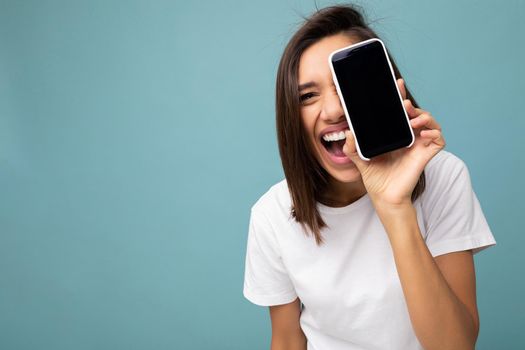 closeup Photo of pretty positive young brunette woman good looking wearing white t-shirt standing isolated on blue background with copy space holding smartphone showing phone in hand with empty screen screen for cutout looking at camera.