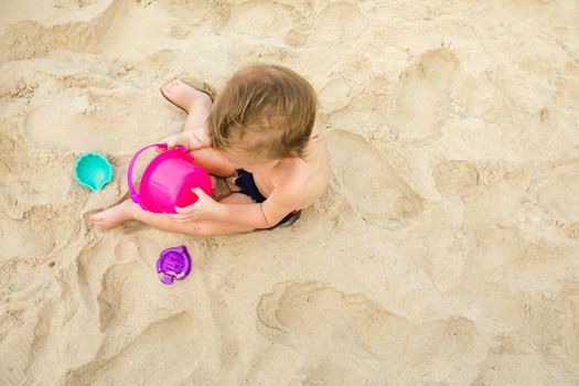 From above shot of child sitting on sand and playing with colorful beach toys.