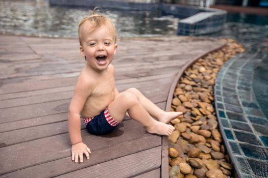 Innocent happy infant sitting on wooden poolside of resort and laughing excitedly.