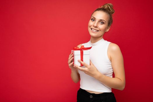 Shot of attractive positive smiling young blonde woman isolated over colourful background wall wearing everyday trendy outfit holding gift box and looking at camera. Empty space