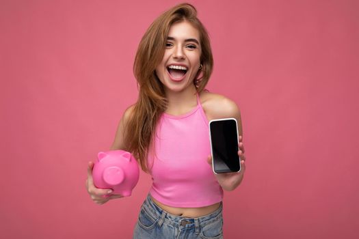 Happy smiling beautiful happy young woman wearing casual outfit isolated over background with empty space holding in hand mobile phone and showing smartphone with empty screen for mockup and money box looking like pink pig looking at camera.