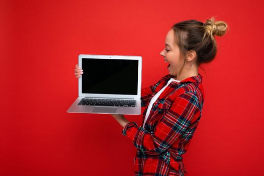 side profile photo of Beautiful amazed young woman holding laptop wearing red shirt looking at monitor display isolated on red background. Mock up