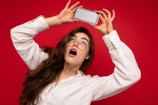 attractive amazed young brunet woman wearing white shirt and optical glasses isolated over red background holding in hand and showing mobile phone with empty screen for mockup looking at gadjet screen.