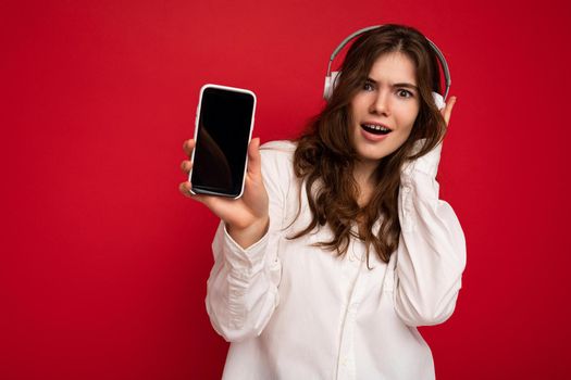 Attractive positive shocked young brunette curly woman wearing white shirt isolated on red background wall holding and showing mobile phone with empty display wearing white bluetooth headphones listening to music looking at camera.