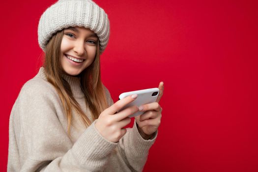 Beautiful sexy smiling young blonde woman wearing casual beige sweater and beige hat isolated over red background holding in hand and using mobile phone communicating online on the internet looking at camera. copy space