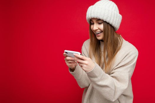 Beautiful young blonde woman wearing casual beige sweater and beige hat isolated over red background holding in hand and using mobile phone communicating online on the internet looking at gadjet display. copy spca