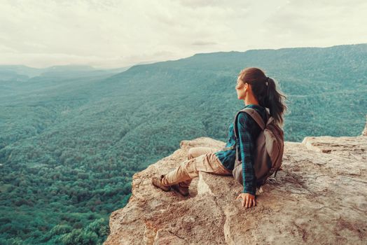 Traveler young woman sitting on edge of cliff and looking on mountains in summer. Toned image