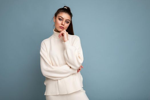Young brunette woman with wearing white casual sweater isolated over blue background looking confident at the camera with crossed arms and hand raised on chin. Thinking positive.